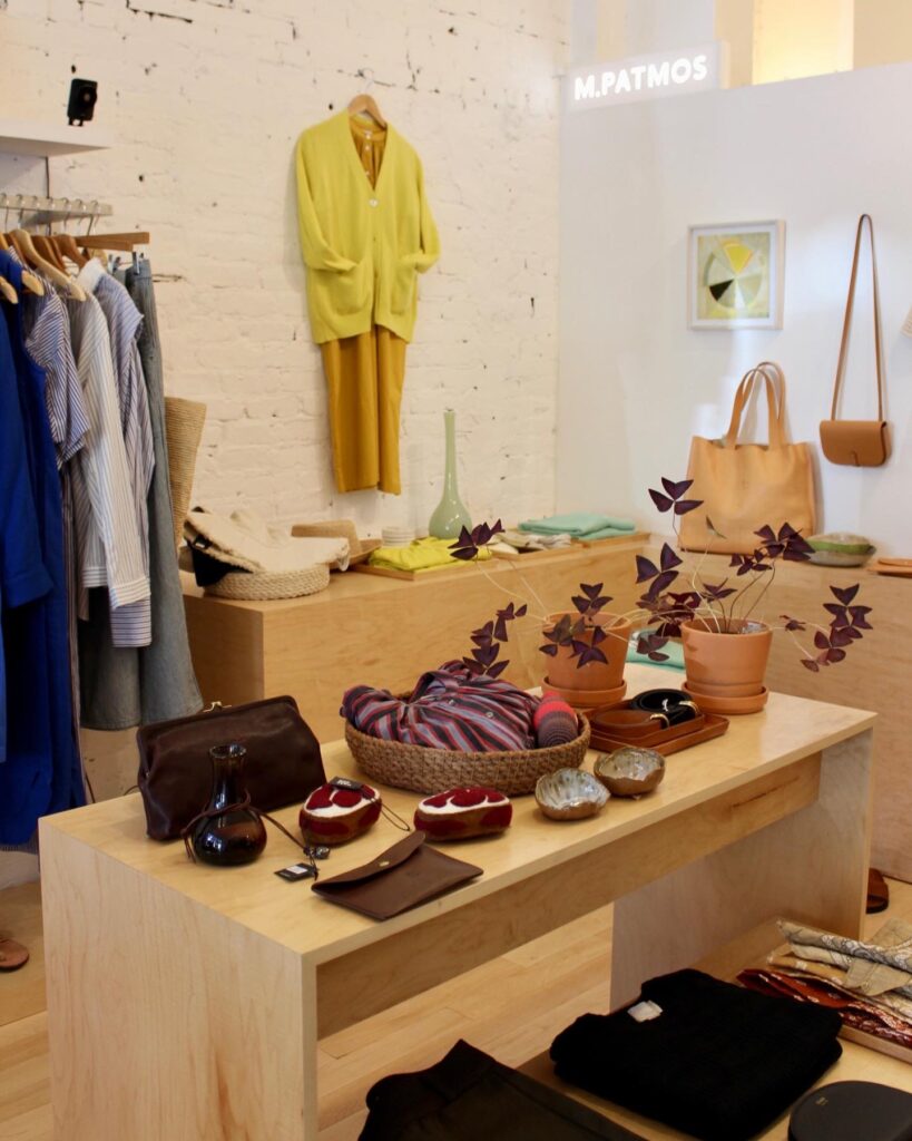 The inside of M Patmos fashion shop in Boerum Hill with a wood table of purses, a rack of clothes to the left, and an outfit hanging on the back wall.