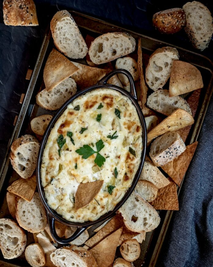 artichoke dip - table magazine Top down view of a cookie sheet filled with various pieces of bread and pita triangles and a warm artichoke dip in the middle.