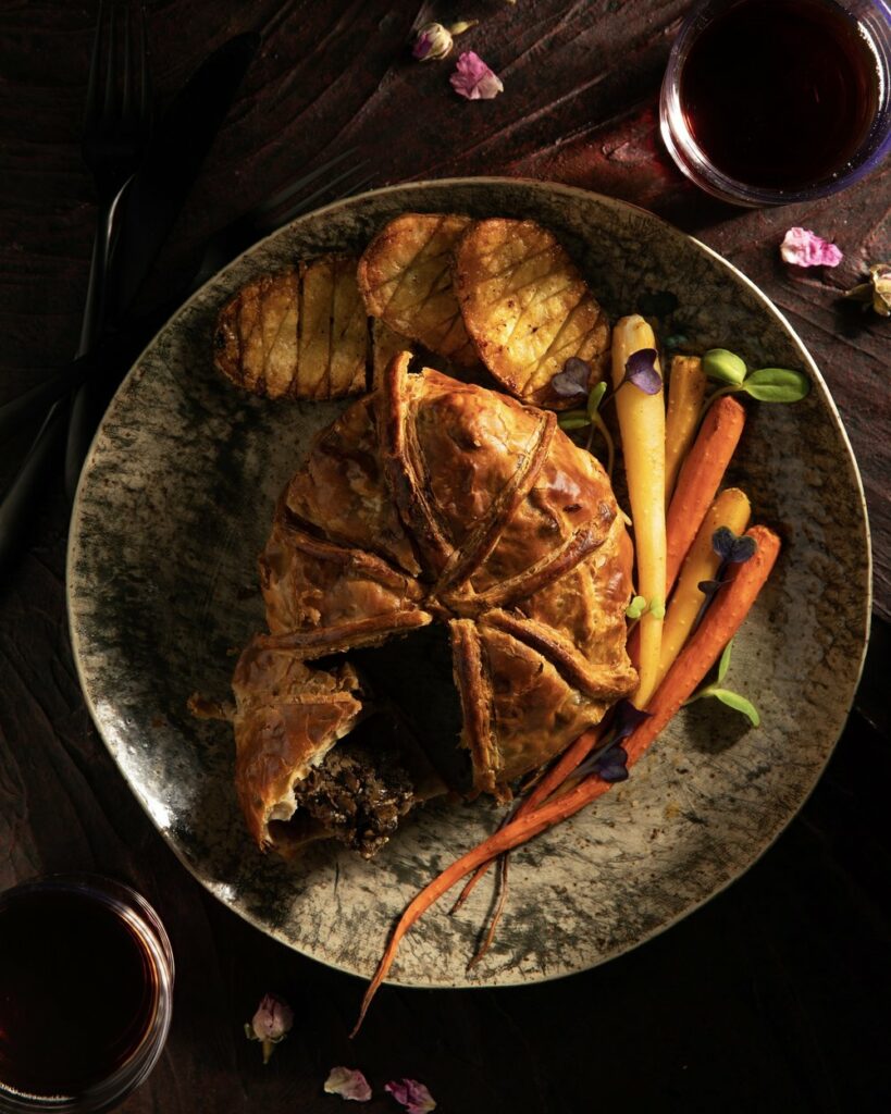 A piece of beef wellington sits cut in teh middle of a plate with roasted potatoes and orange and yellow carrots.