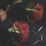 A beet-colored bloody mary on a black backgroound, garnished with a sprig of rosemary.
