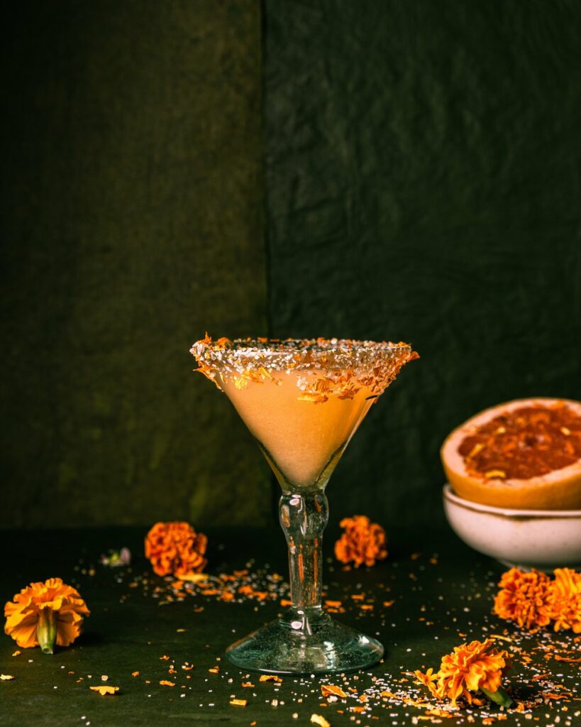 An orange cocktail in a cocktail glass with marigolds all around the table and a grapefruit in the background.