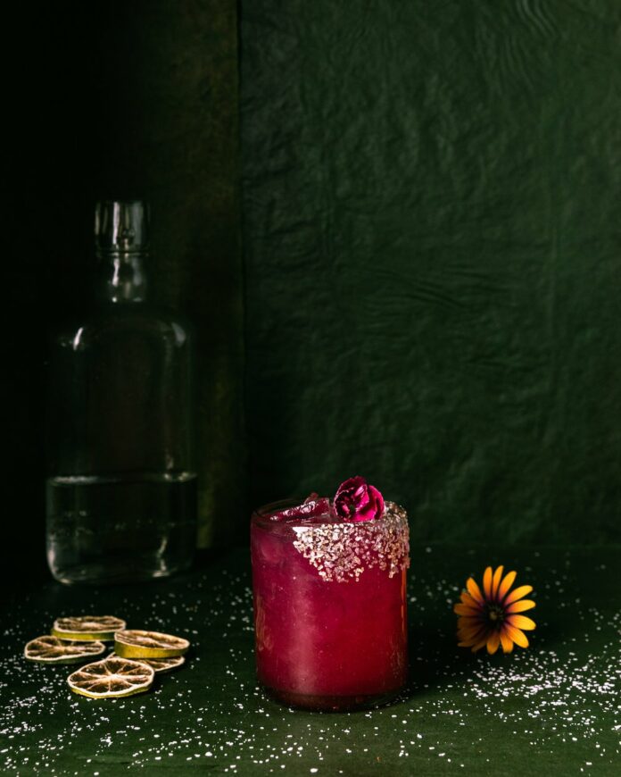 Paloma's+Prickly+Pear+Margarita+TABLE+Magazine+Howard+2 A dark-pinkish red prickly pear margarita sits in a salt-rimmed glass in front of a green background.
