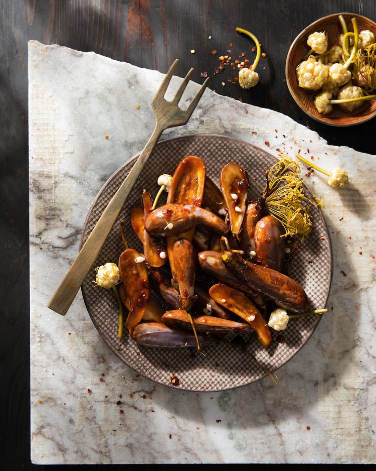 A brown plate of sticky eggplant with pickled garlic blossoms throughout as a big fork sits over top of the bowl.