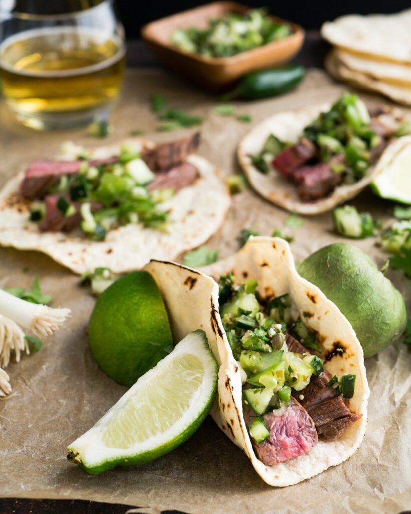 Flank steak tacos in a tortilla topped in a roasted pepper salsa beside slices of lime.
