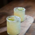 Two glasses with a pale yellow margarita with lime slices floating on top.