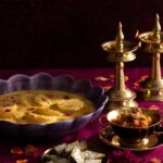 A dark background with a deep, rich red surface where a purple scalloped shallow bowl holds a Diwali dessert next to gold candle sticks.