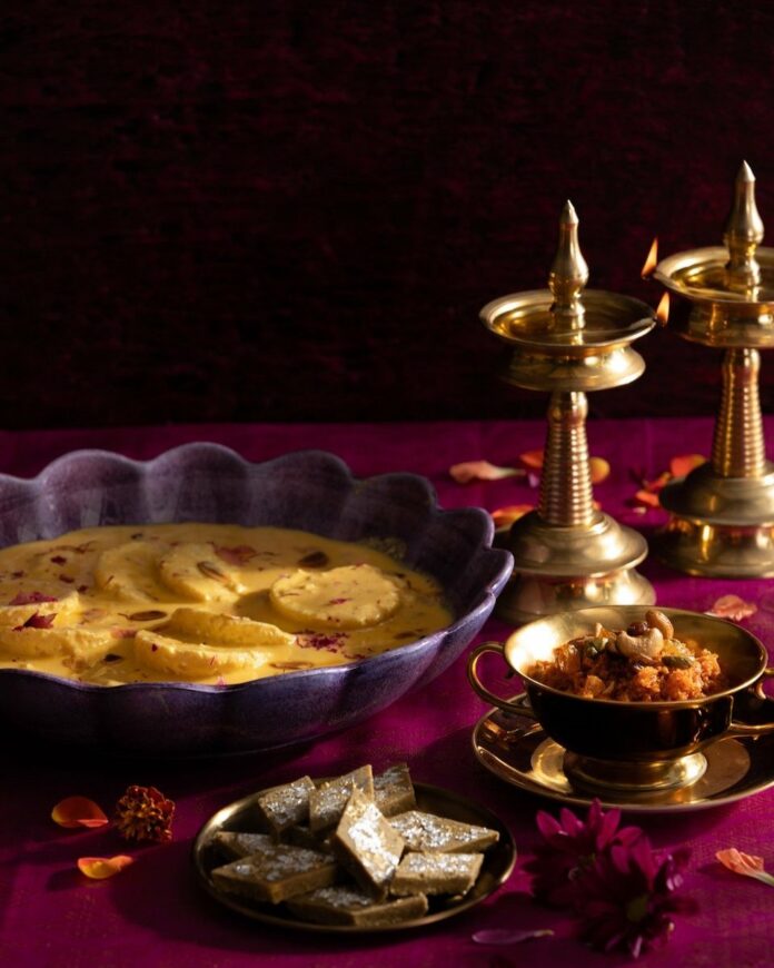 A dark background with a deep, rich red surface where a purple scalloped shallow bowl holds a Diwali dessert next to gold candle sticks.