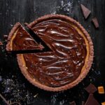 Layers of Earl Grey caramel filling and rich chocolate ganache nested into a cocoa crust make up this delectable chocolate tart. On a black background sits the chocolate tart with a single triangle slice cut out.