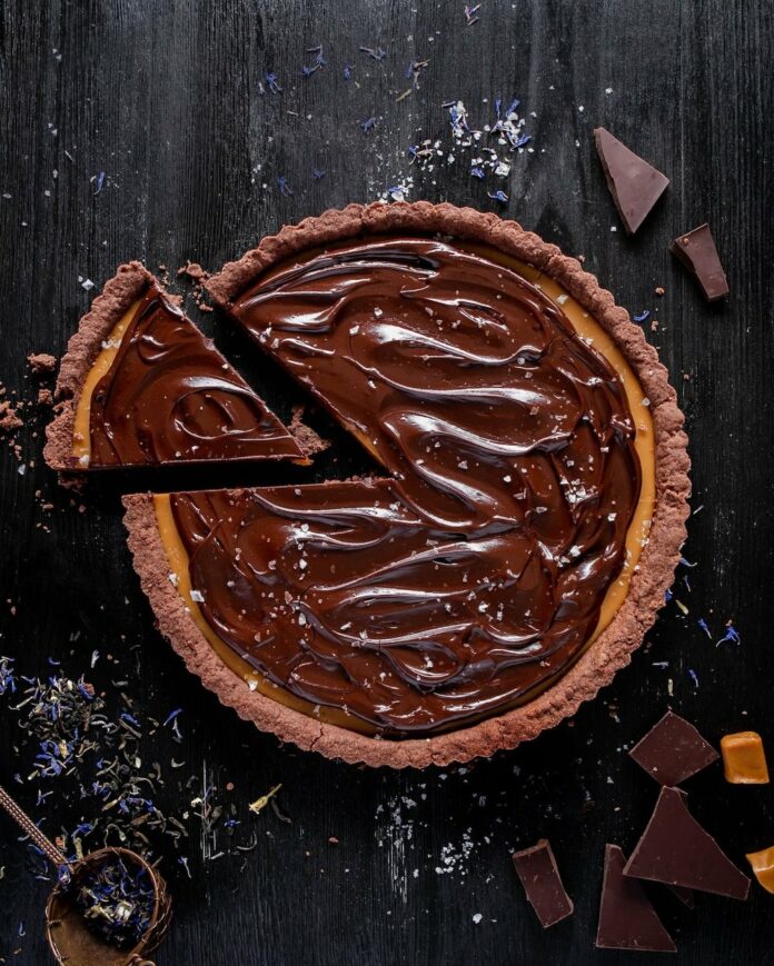 Layers of Earl Grey caramel filling and rich chocolate ganache nested into a cocoa crust make up this delectable chocolate tart. On a black background sits the chocolate tart with a single triangle slice cut out.