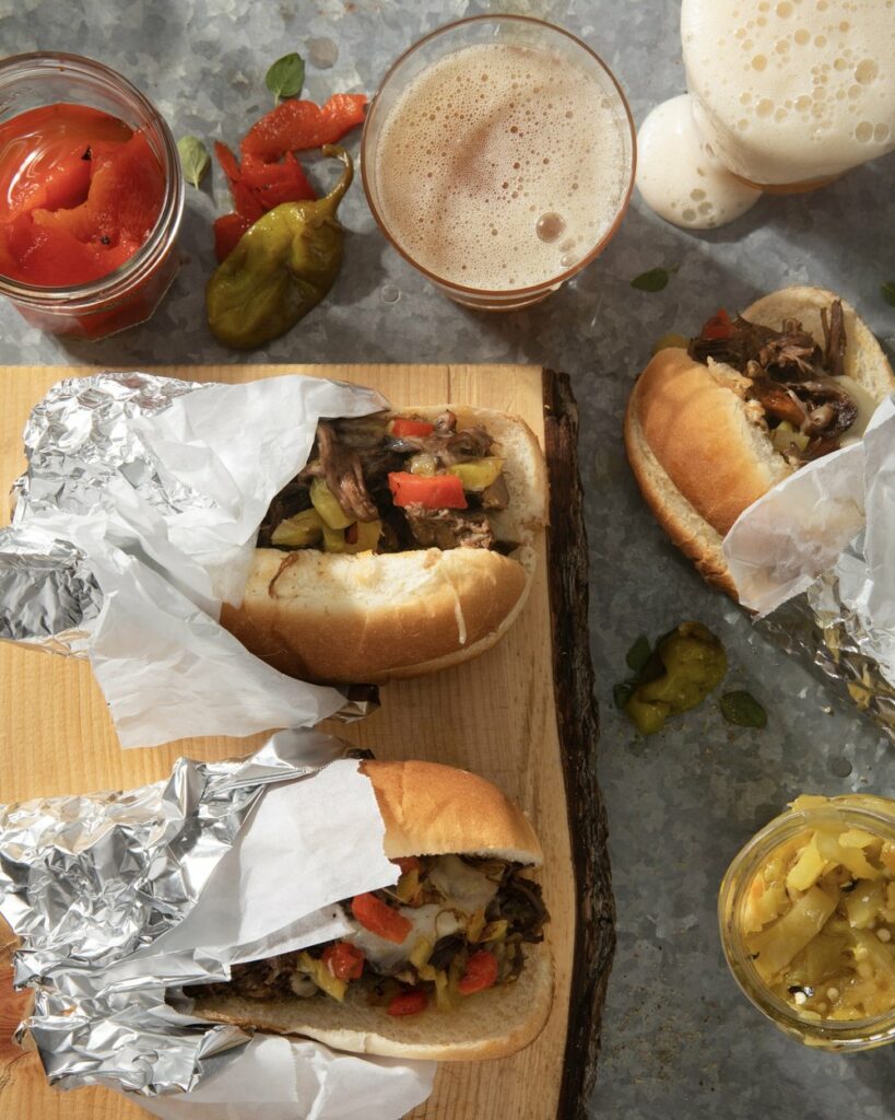 Three Italian Beef Sandwiches lay in various directions surrounded by fried peppers.