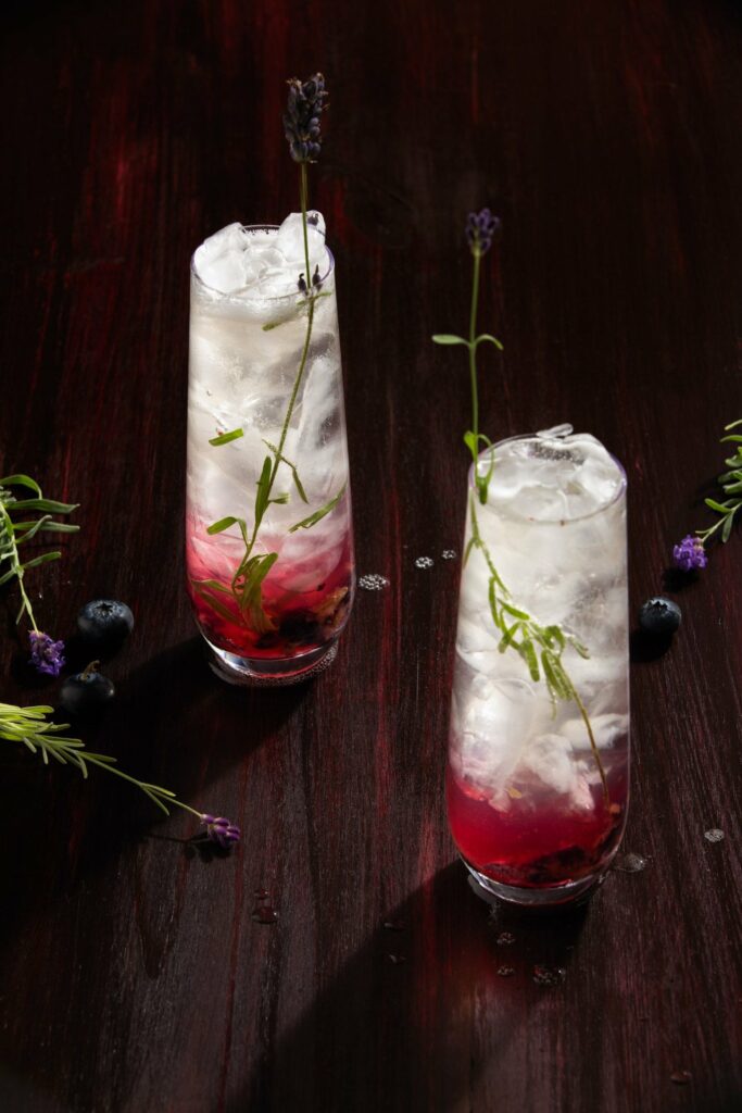 A lavender mocktail in a long, tall glass with maroon and lavender colors and a sprig or rosemary for garnish