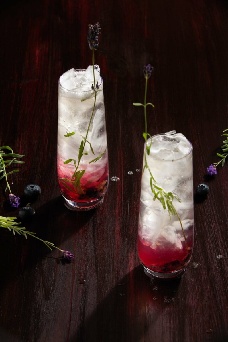 A lavender mocktail in a long, tall glass with maroon and lavender colors and a sprig or rosemary for garnish 