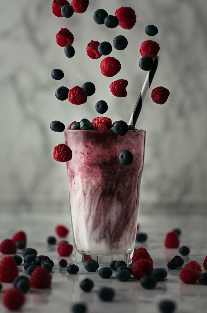 Blueberrys and raspberries fall into a milkshake glass filled with a white milkshake with the aforementioned berries.