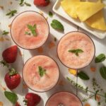 Three glasses of strawberry pineapple agua fresca sit in glasses on a white picnic table surrounded by strawberries and slices of pineapple.