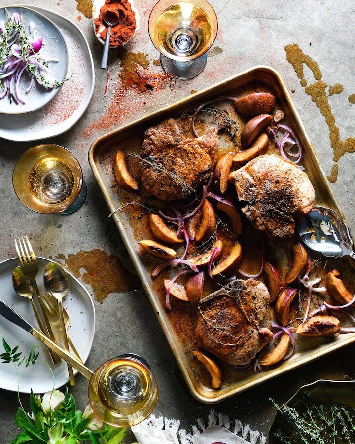Pork chops and peaches sit in a sheet pan with plates and glasses nearby.