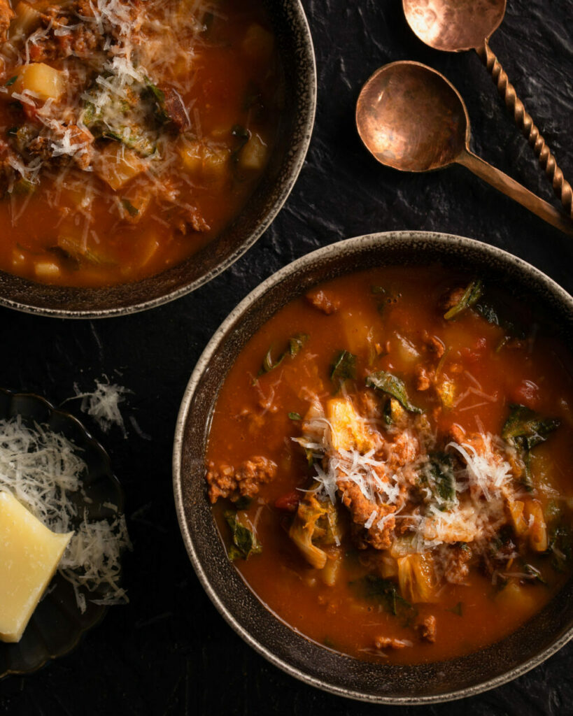 Two bowls filled with a dark reddish brown broth with spicy sausage and greens as butter sits to the left and two spoons sit in the top right.