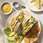 A turkey and apple sandwich side up on a white plate next to chips.