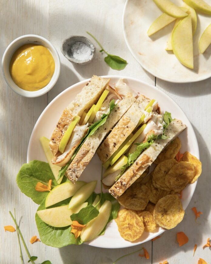 A turkey and apple sandwich side up on a white plate next to chips.