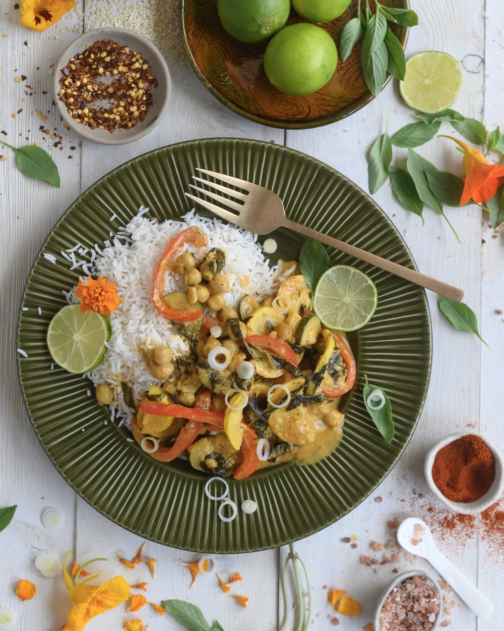 Vegan squash curry with rice garnished with lime and fresh herbs on a green plate, surrounded by fresh ingredients and spices.