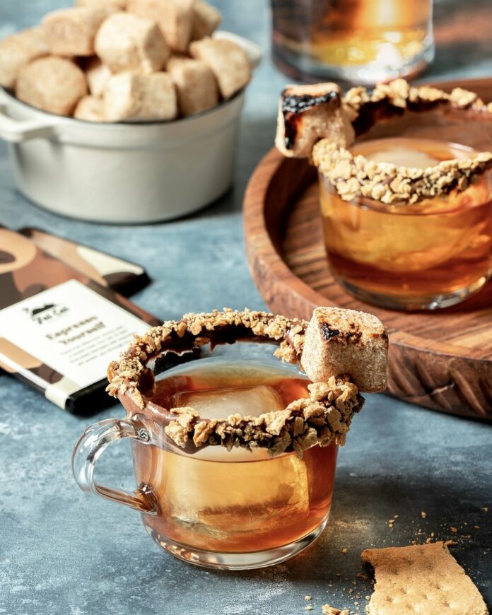 A chocolate-hazelnut spread mixed with crushed graham cracker rim two glasses filled with a whiskey cocktail, garnished with a roasted marshmallow.