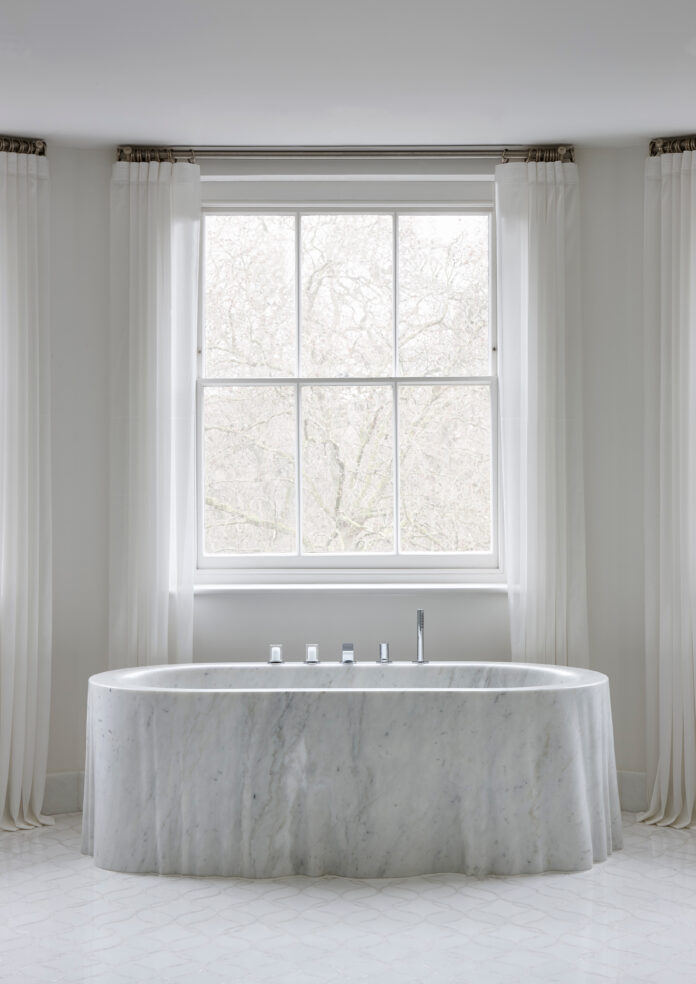 A photo of a marble bath tub in a pale gray room in front of a large window.