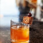 An orange in color old fashioned cocktail sits in a old fashioned glass, garnished with banana bread.