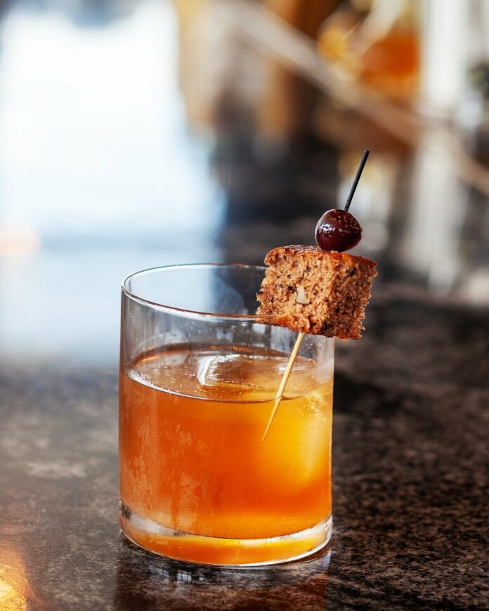An orange in color old fashioned cocktail sits in a old fashioned glass, garnished with banana bread.
