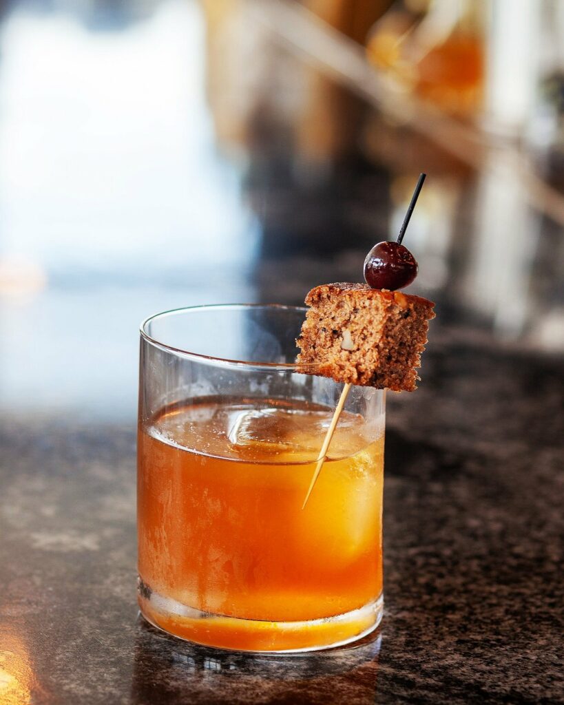 An orange in color old fashioned cocktail sits in a old fashioned glass, garnished with banana bread.