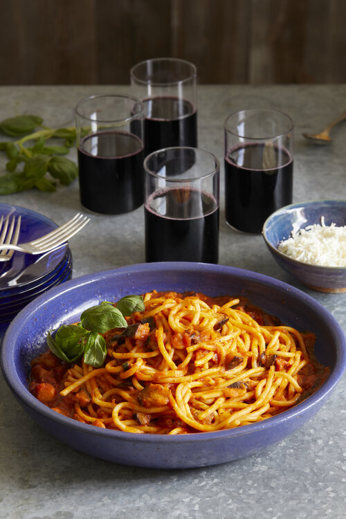 A Stanley Tucci Inspired Bucatini all' Amatriciana sits in a blue bowl with four glasses of wine placed behind the pasta.