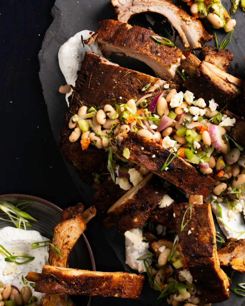  A board with sliced baby back ribs covered with a white bean salad and a white sauce on a black surface. Tailgating Recipes