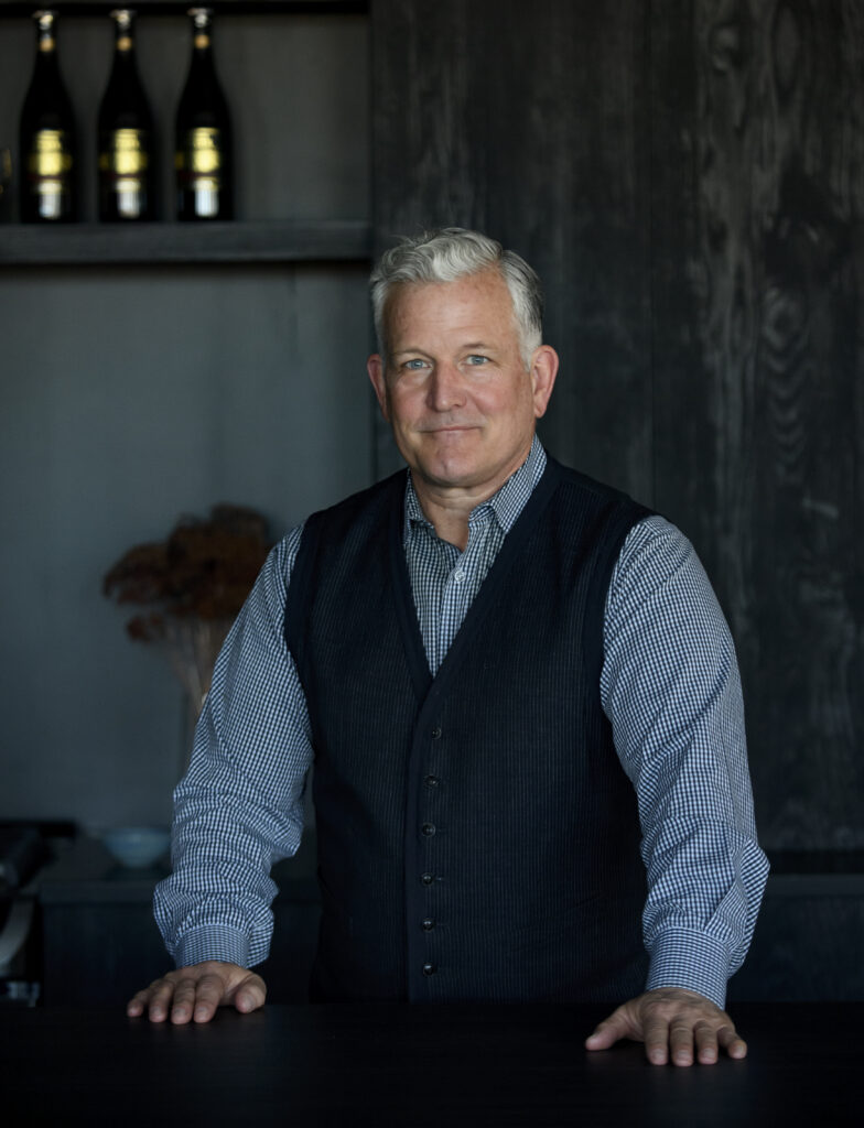 Keith Recker stands at a counter in a black vest and blue button down. 