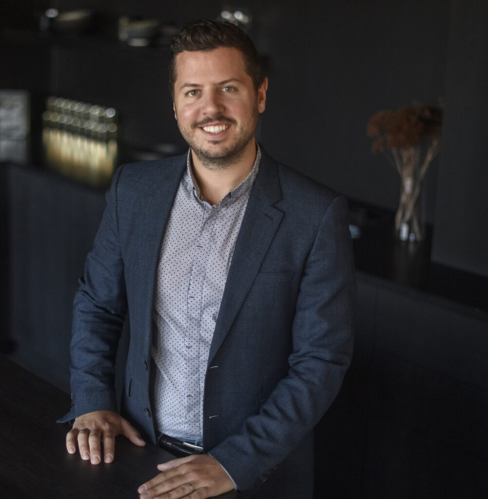Justin matase stands at a counter top in a button up and suit jacket. 