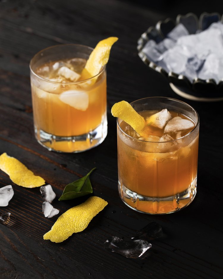 2 identical whisky cocktails in rocks glasses, garnished with lemon peel with a black background