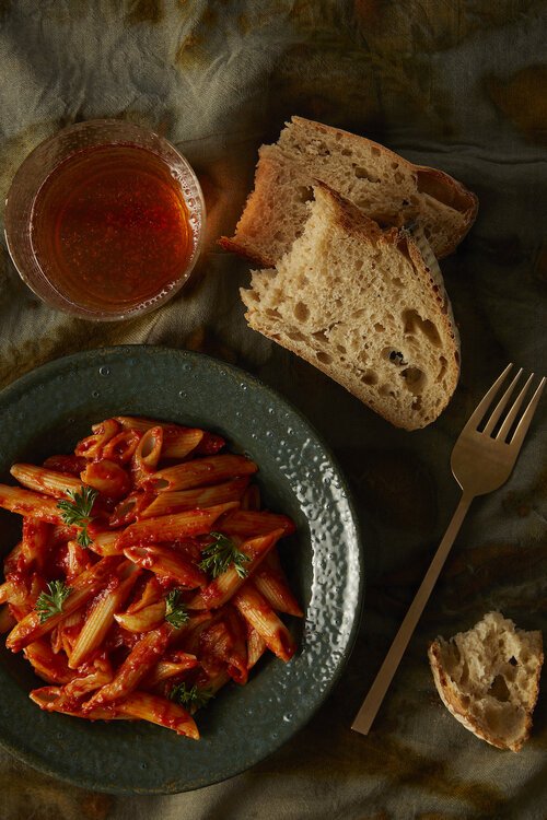 Stanley Tucci Inspired Penne all’ Arrabbiata sits in a green ceramic bowl with two slices of french bread off to the right.