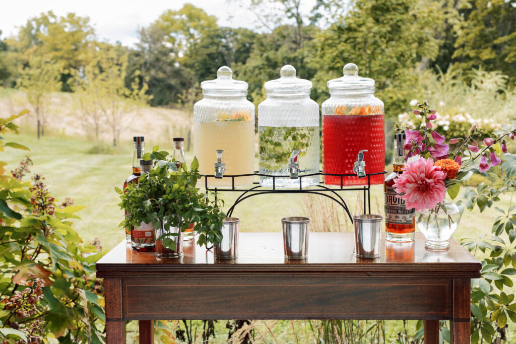 Three glass beverage dispensers with yellow, clear, and red drinks on a wooden table with flowers set up outside