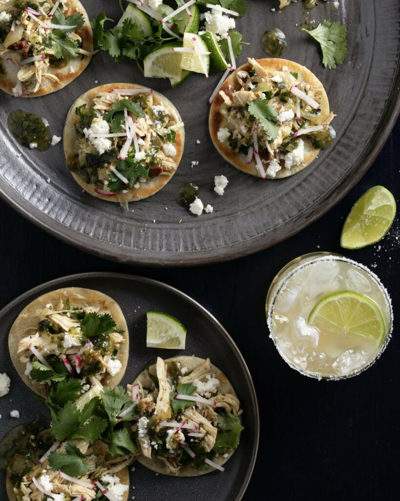 Mini chicken tostados on 2 different plates with a black surface and a margarita on the side.