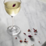 A slightly yellow in color Rose and Black Pepper Martini in a martini glass sits on a white granite countertop. Rose petals are off to the side.