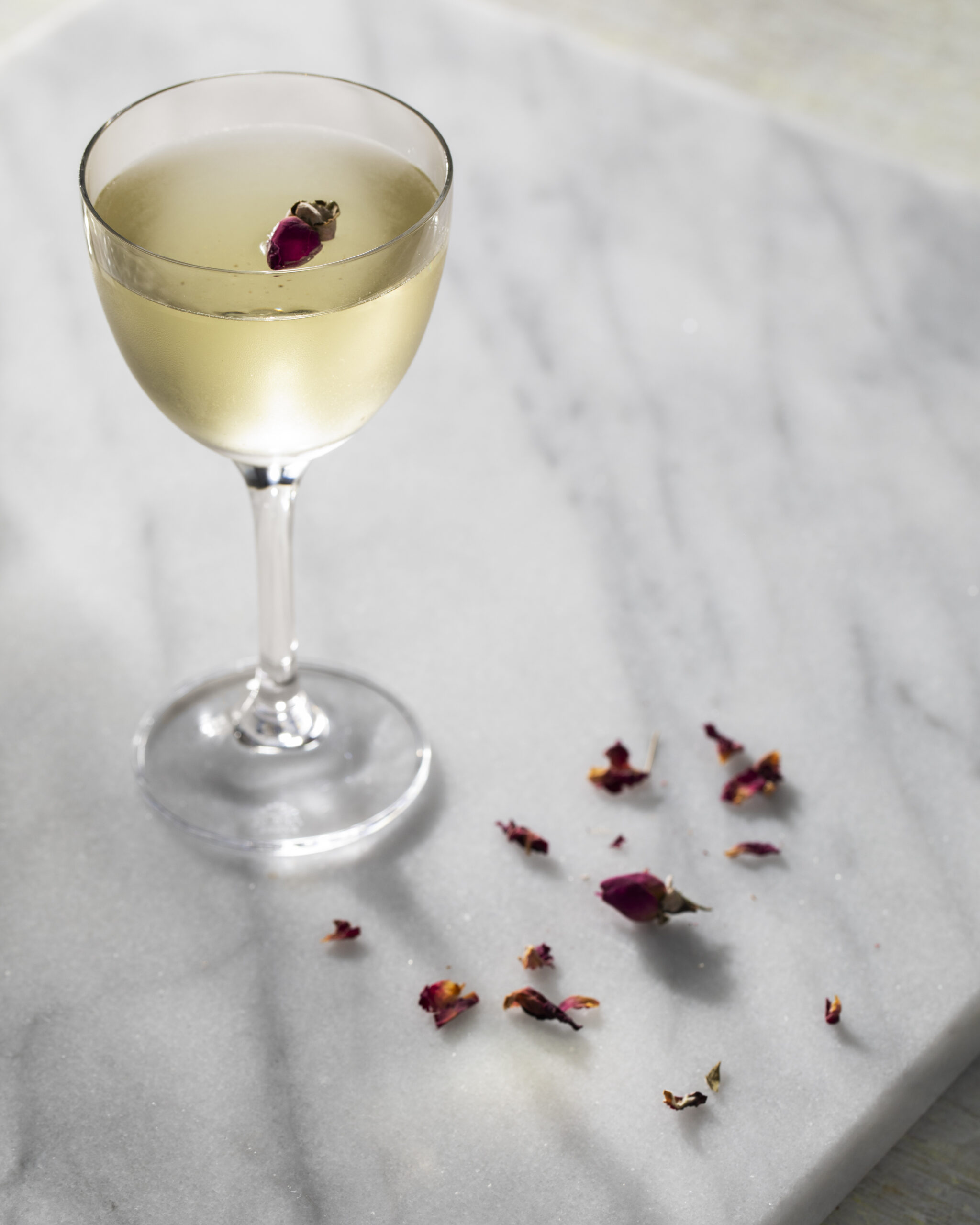 A slightly yellow in color Rose and Black Pepper Martini in a martini glass sits on a white granite countertop. Rose petals are off to the side.