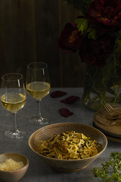 A bowl of Stanley Tucci Inspired Spaghetti alla Nerano sits next to two glasses of white wine.
