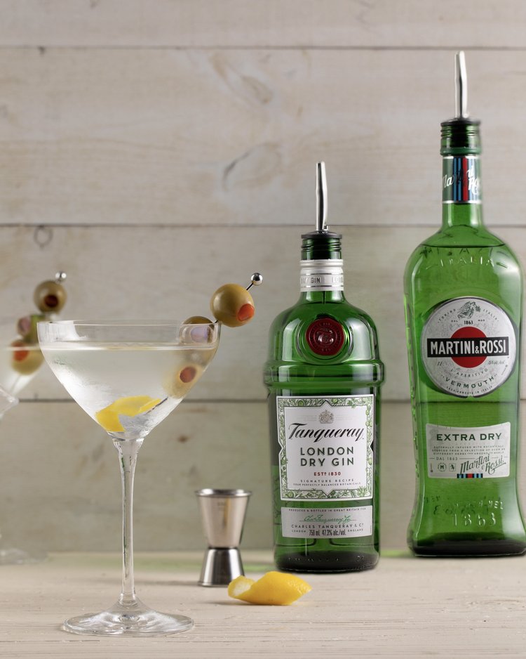 A martini sits beside two green bottles of dry gin.