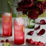 Two skinny glasses of our Hibiscus and Mint Spritz cocktail sit to the left next two a vase of full, blooming, dark red flowers.