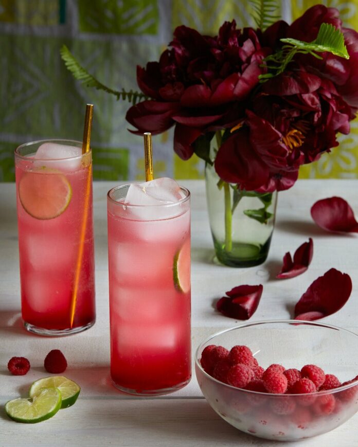 Two skinny glasses of our Hibiscus and Mint Spritz cocktail sit to the left next two a vase of full, blooming, dark red flowers.