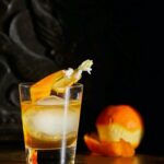 A orange with half its peel off sits to the right of a smoked bourbon old fashioned in rocks glass. curls of smoke waft off the glass.