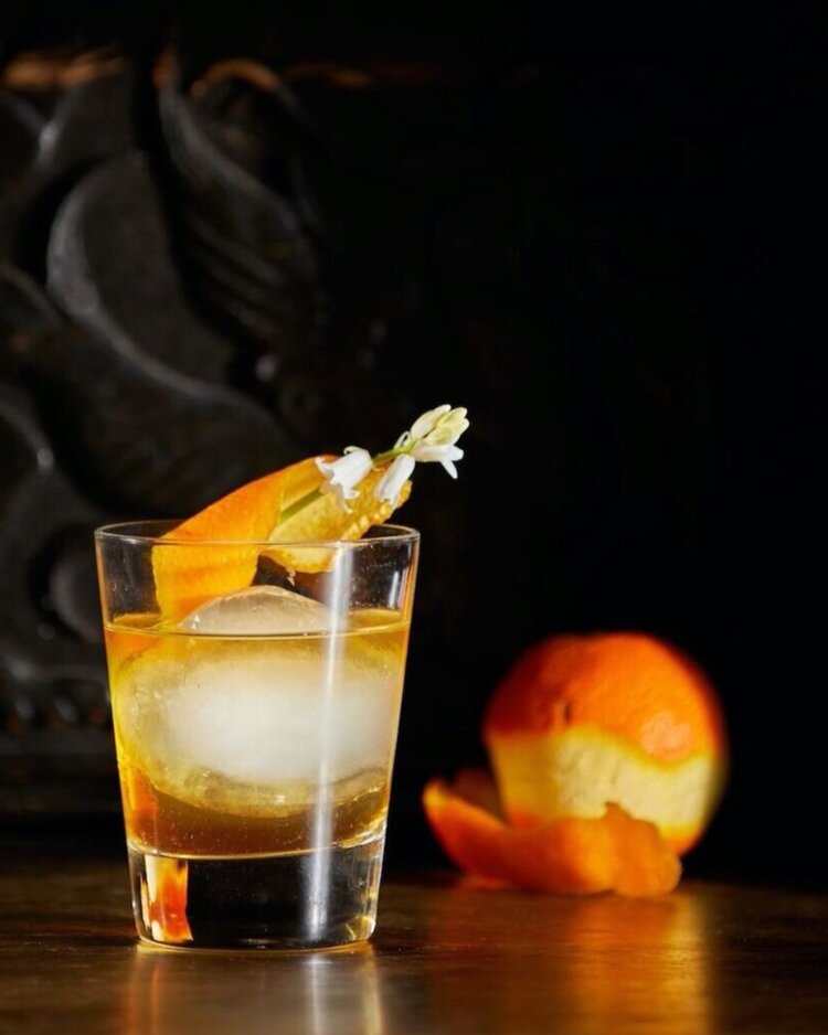 A orange with half its peel off sits to the right of a smoked bourbon old fashioned in rocks glass. curls of smoke waft off the glass.