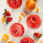 Glasses of a red cocktail garnished with a blood orange slice, surrounded by strawberries, oranges, and mandarin orange slices on a white surface.