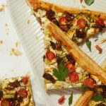 A flat lay photo of several slices of a savory artichoke puff pastry tart, topped with sun-dried tomatoes, feta cheese, and parsley.