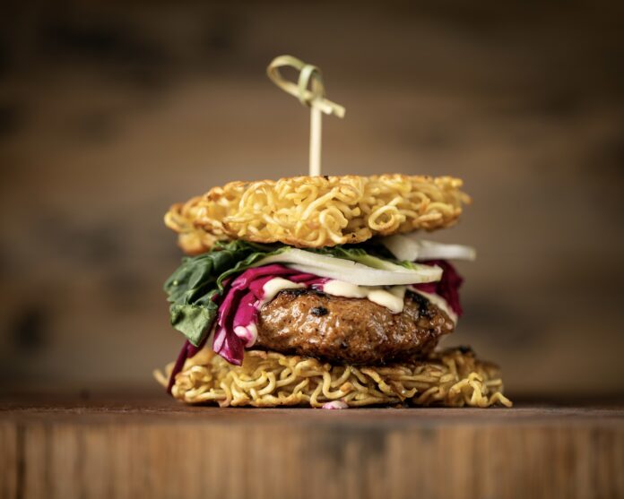 a hamburger with fixings on a bun made of ramen noodles with a bamboo pick holding it all together