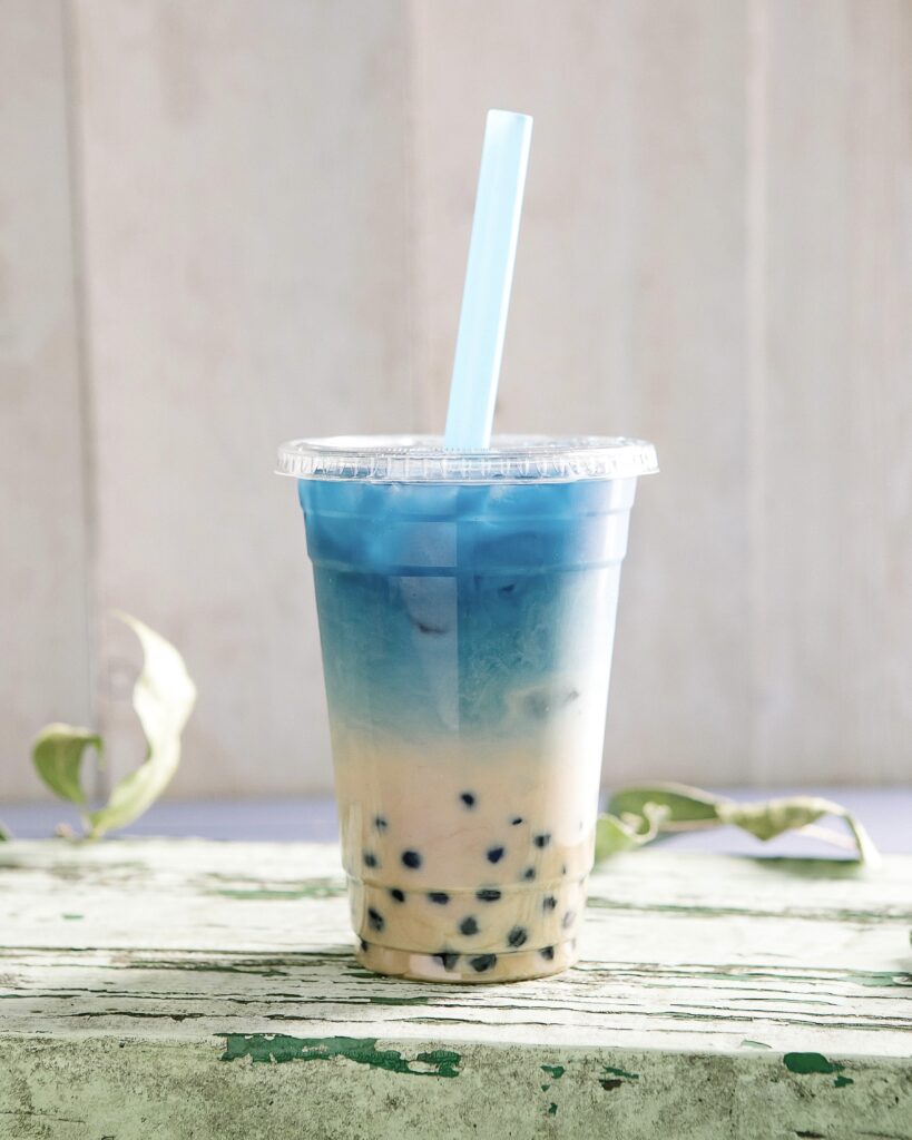 a look in photo with a rustic green table and a white paneled background of a to-go cup with boba tea that is a light tan color at the bottom with dark brown boba and a bright blue butterfly pea tea at the top, a blue straw