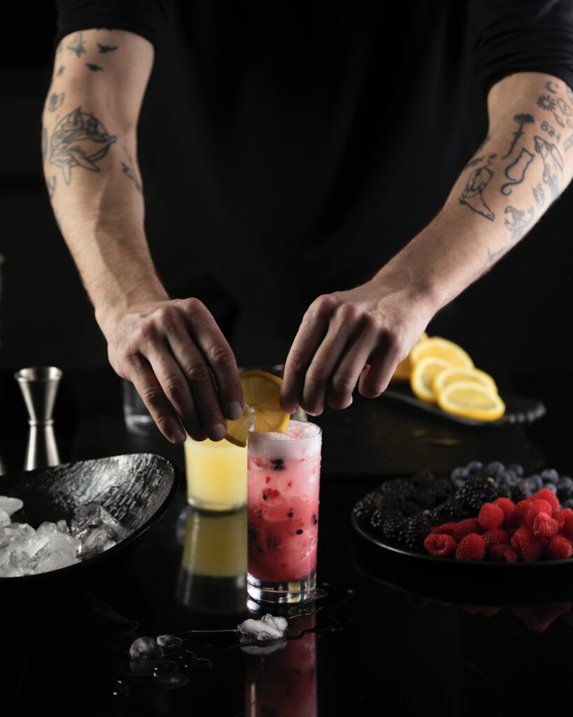a man with tattooed arms garnishing a red mocktail with a lemon slice