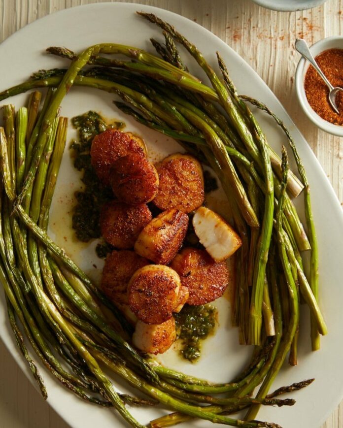 Brown Butter Scallops on a tray with asparagus in the surroundings. A red chili powder in a small bowl with a spoon on the side.